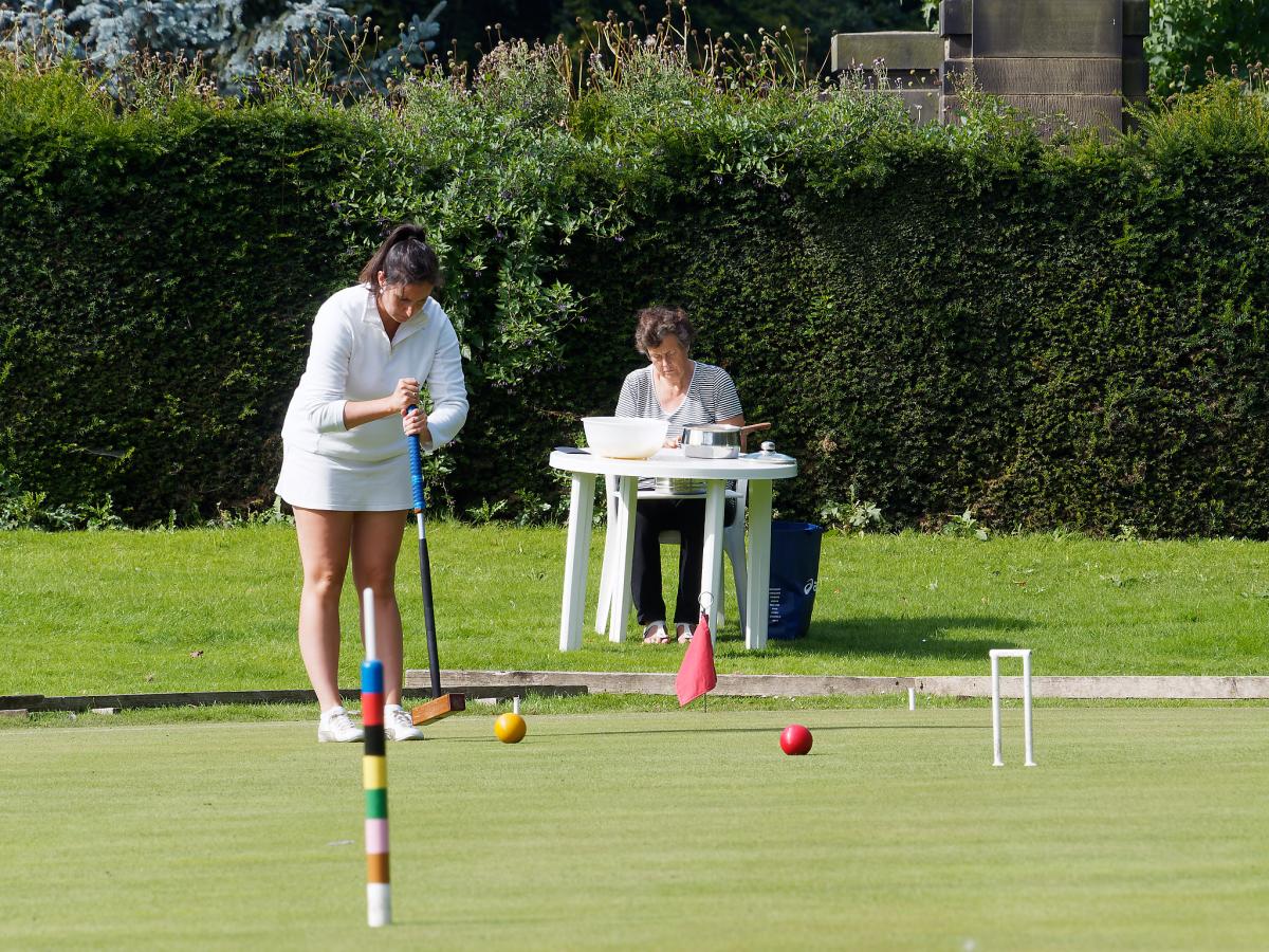 Young woman in whites playing while her mother is sat at a picnic table off court peeling potatoes