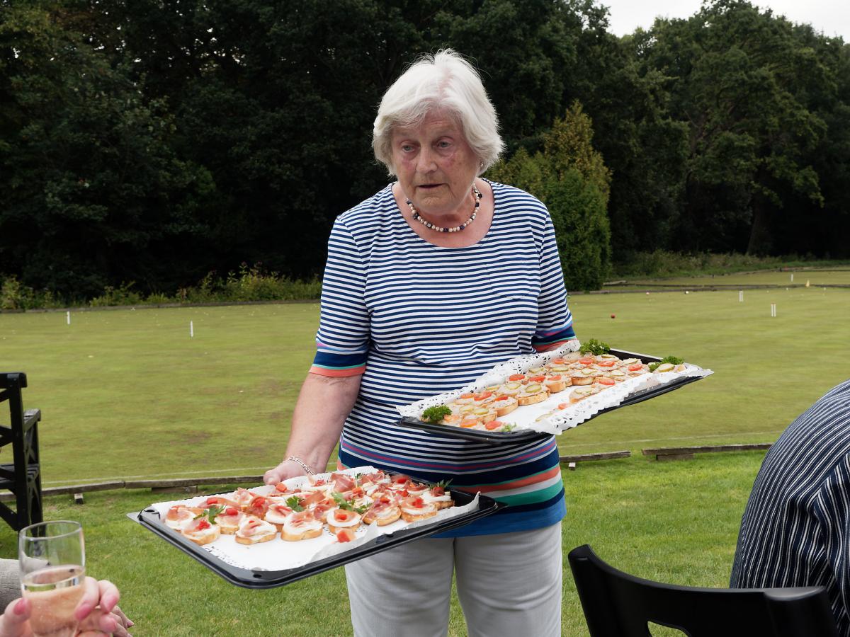 white haired woman offering two large trays of canapes to a person out of shot