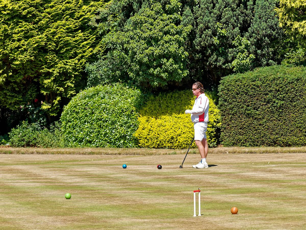 Male player in England strip (white with red flashes) playing on a very browned turf with elegant conifers in the background